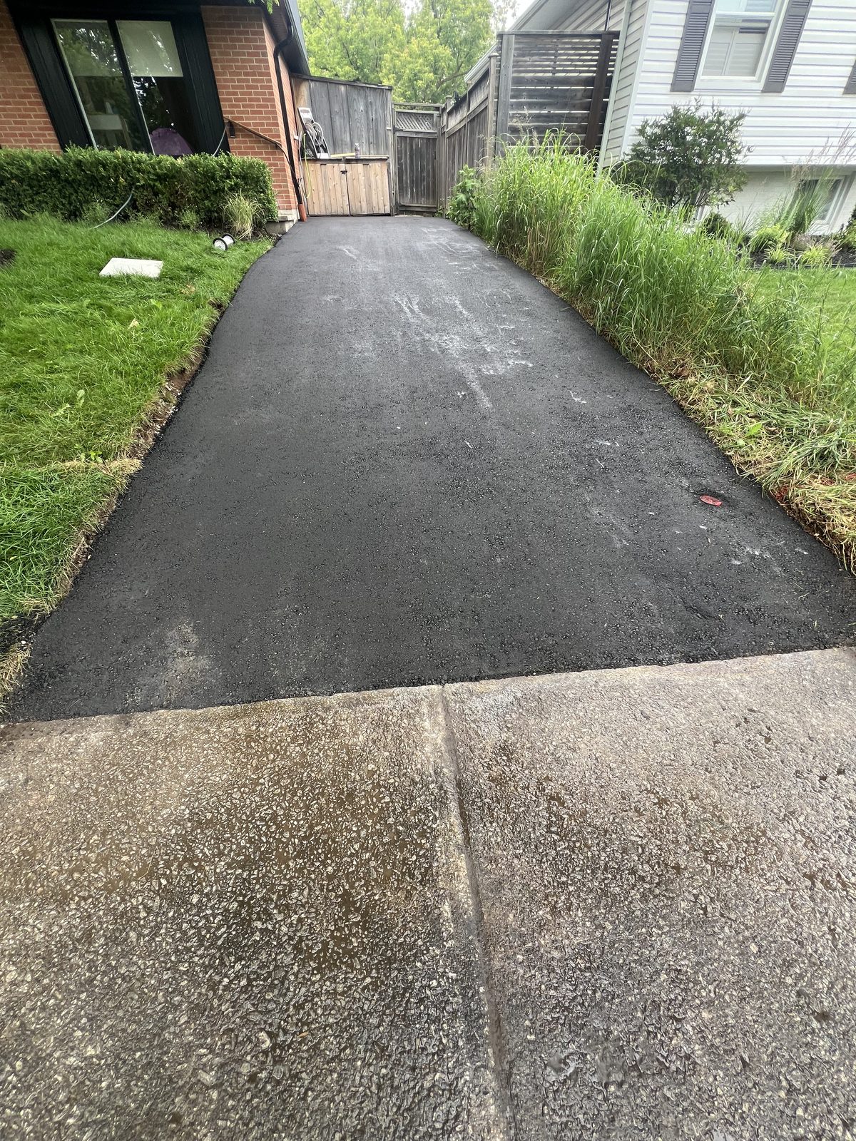 Newly paved smooth asphalt driveway between a brick home and fence in the GTA, meeting an existing concrete apron