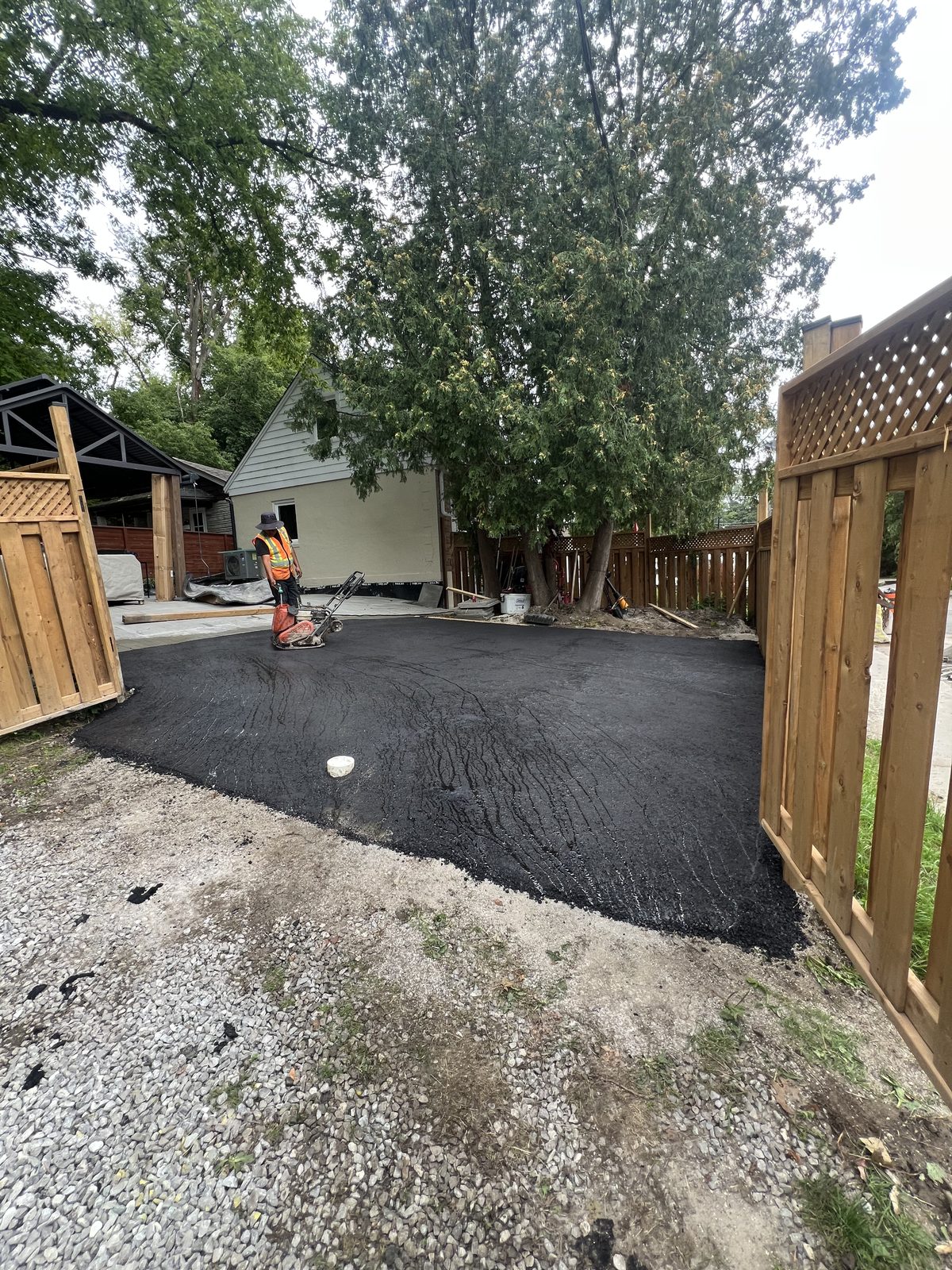 Worker using a plate compactor on a freshly laid asphalt backyard driveway surrounded by wood fencing in the GTA
